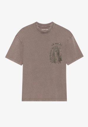 T-shirt oversize in cotone marrone con stampa grafica verde di una figura con fiori e testo sulla parte anteriore, maniche corte e colletto rotondo.