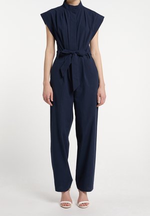 Femme portant une combinaison sans manches bleu marine avec un col montant et une ceinture nouée à la taille, associée à des sandales blanches à bouts ouverts, debout devant un fond uni.