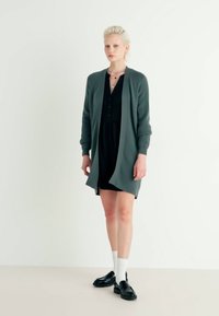 Long grey cardigan made of soft fabric, open front, ribbed cuffs, layered over a black dress. Model wears black shoes and white socks.