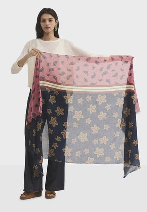 Žena u bijeloj majici i tamnim hlačama drži veliki pravokutni šal s ružičastim i tamnoplavim cvjetnim i paisley узорима.