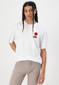 White cotton t-shirt featuring a red circle graphic and black text. Short sleeves, relaxed fit, and a smooth texture. Model wearing beige pants.
