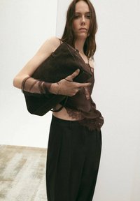 Mujer con blusa marrón translúcida con encaje y pantalones negros, sosteniendo un gran bolso oscuro de textura sobre un fondo blanco.