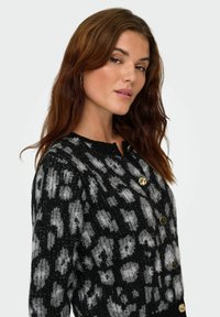 Femme avec des cheveux bruns ondulés portant un cardigan boutonné noir et gris à motifs, regardant légèrement par-dessus son épaule vers l'appareil photo.