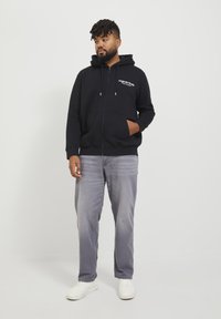 Sort zip-up hoodie med hvid tekst, kængurulomme og snore. Parret med lysegrå jeans og hvide sneakers. Ensfarvet baggrund.