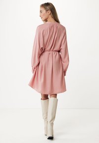 Abito rosa a maniche lunghe con vestibilità comoda, scollatura arricciata e cintura abbinata. Indossato con stivali bianchi al ginocchio su uno sfondo neutro.