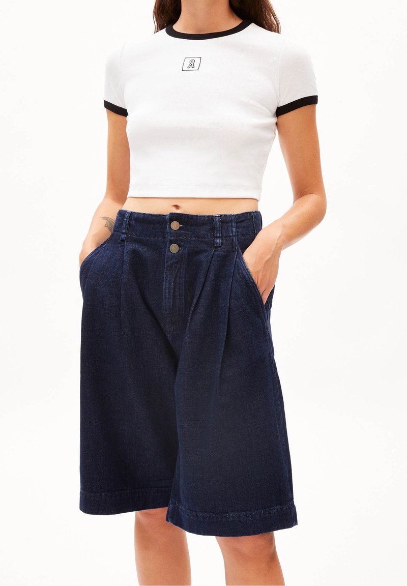 Femme portant un t-shirt court blanc avec un bord noir et un short en denim bleu foncé taille haute à jambes larges avec des poches.