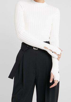 Osoba ubrana w biały prążkowany długi rękaw z otworami na kciuki oraz czarne spodnie z wysokim stanem, plisowane, z paskiem.