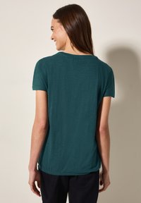 T-shirt teal a maniche corte in un fit rilassato, con scollatura rotonda e una texture sottile. Il retro è leggermente più lungo con orlo arrotondato.