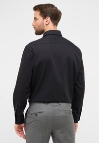 Maglietta nera a maniche lunghe con colletto standard, abbinata a pantaloni grigi con tasche con zip e finitura testurizzata.
