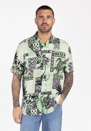 Mężczyzna z tatuażami, ubrany w zielono-czarną hawajską koszulę z krótkim rękawem i niebieskie dżinsy, stojący na białym tle.