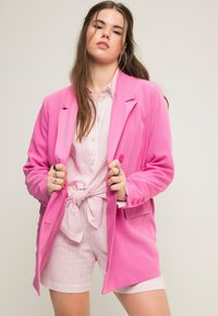 Blazer rosa con hombros estructurados, diseño de un solo botón y detalles de botón. Llevarlo sobre una camisa a rayas y pantalones cortos a juego.