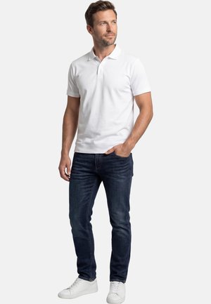 Mann steht mit einer Hand in der Tasche, trägt ein weißes Poloshirt, dunkle Jeans und weiße Turnschuhe vor einem schlichten Hintergrund.