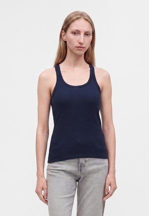 Jonge vrouw met lang blond haar, gekleed in een marineblauw mouwloos shirt en lichtgrijze spijkerbroek tegen een effen achtergrond.