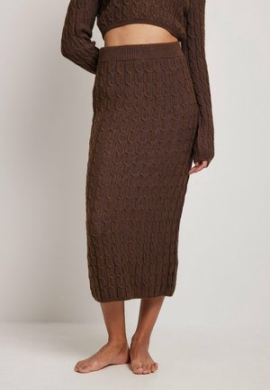 Femme portant une jupe midi marron en maille torsadée et un pull court assorti, debout pieds nus devant un fond uni.