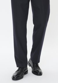 Chaussures en cuir noir poli assorties à un pantalon bleu marine. Le pantalon présente un tissu texturé ; les chaussures ont des bouts ronds et des lacets.