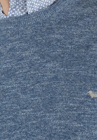 Pull en tricot bleu avec un motif texturé, comprenant un col rond et un petit logo beige en accent. Matière douce de poids moyen.