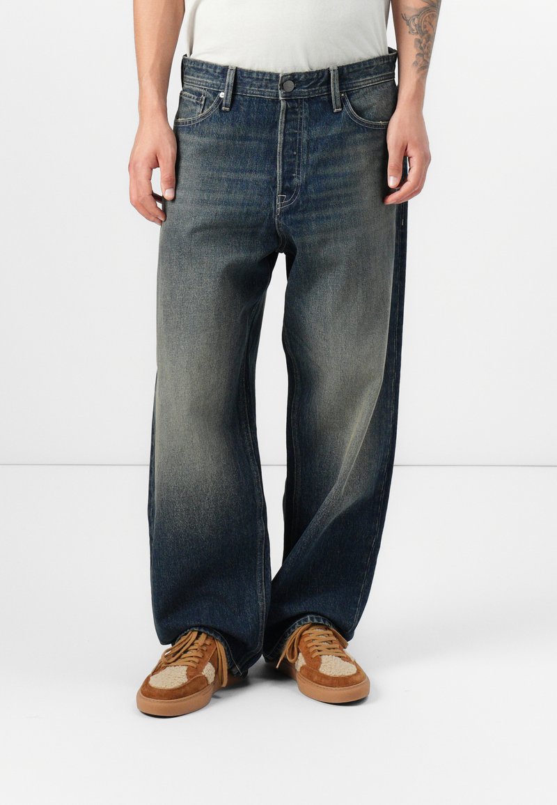 Wijde donkerblauwe spijkerbroek van denim met een vervaagde was, vijf zakken en metalen accessoires; gecombineerd met bruine en beige sneakers.