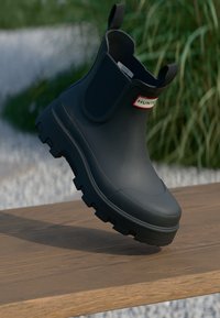 Bota de tobillo de goma negra con suela texturizada, que cuenta con una etiqueta roja y blanca en el lateral. Acabado suave y diseño tipo slip-on.