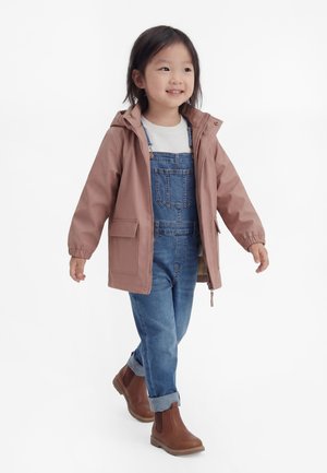 Copil zâmbitor care merge, purtând o geacă roz cu glugă, cămașă albă, salopetă din denim albastru și ghete maro până la gleznă pe fundal alb.