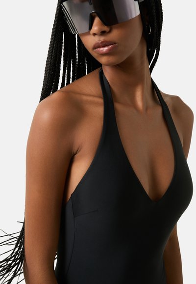 Black halter neck swimsuit with a deep V-neck and smooth fabric, featuring a subtle side seam detail. Accompanied by oversized sunglasses.