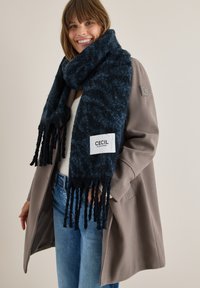 Mørkeblå tørklæde med struktureret overflade, frynser i kanten og et hvidt mærke med teksten "CECIL", bæres over en lys sweater og en beige frakke.
