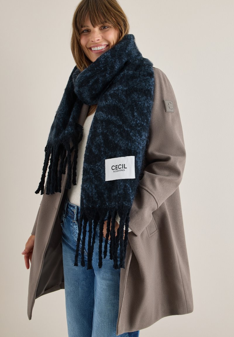 Scarf blu scuro con finitura strutturata, bordi frangiati e un'etichetta bianca che recita "CECIL", indossato sopra un maglione chiaro e un cappotto beige.