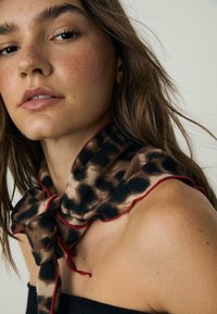 Løveprint tørklæde med røde kanter, lavet af blødt stof, bundet om halsen. Har et lysebrunt og sort mønster på en beige baggrund.