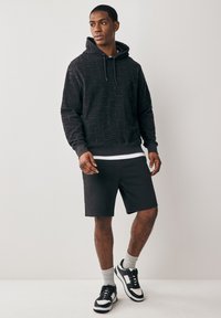 Svart hoodie med texturerat geometriskt mönster, känguruficka och justerbar dragsko. Kombinerad med svarta shorts och sneakers med vita detaljer.