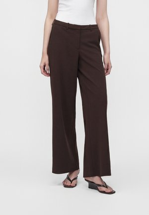 Femme portant un pantalon large marron foncé, un haut blanc sans manches et des sandales compensées noires, debout devant un fond uni.