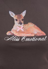 Noor vasikas roosa paelaga kaela ümber, lebamas tume kangapõhja peal kursiivkirjas tekst "Miss Emotional" kohal.