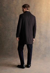 Czarny wełniany płaszcz o dopasowanym kroju, nieco dłuższej długości, z detalami w postaci guzików na rękawach, zestawiony z ciemnymi dżinsami i czarnymi botkami.