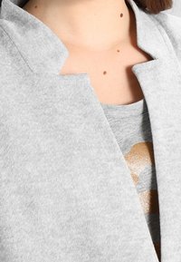 Zbliżenie na osobę noszącą jasnoszarą otwartą kurtkę na szarą koszulę z częściowym złotym nadrukiem, ukazującą pieprzyki na szyi i klatce piersiowej.