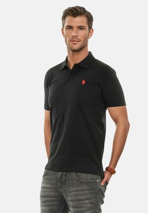 Black short-sleeve polo shirt with a collar, featuring a small red logo on the chest. Worn with gray jeans; smooth fabric texture.