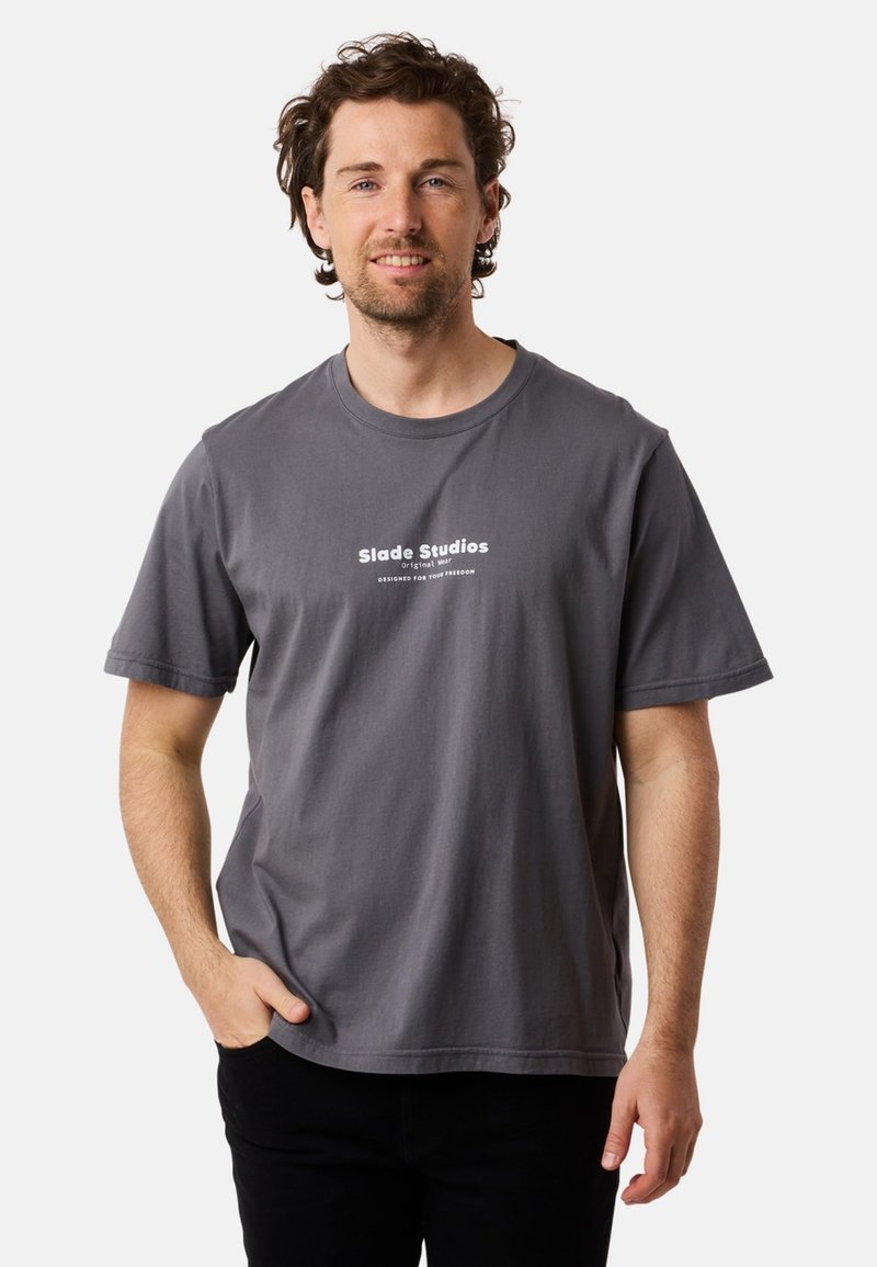 Harmaa puuvillapaita lyhyillä hihoilla, pyöreällä kaula-aukolla, ja etupuolella tuloste, jossa lukee "Slade Studios Original Wear" valkoisella.