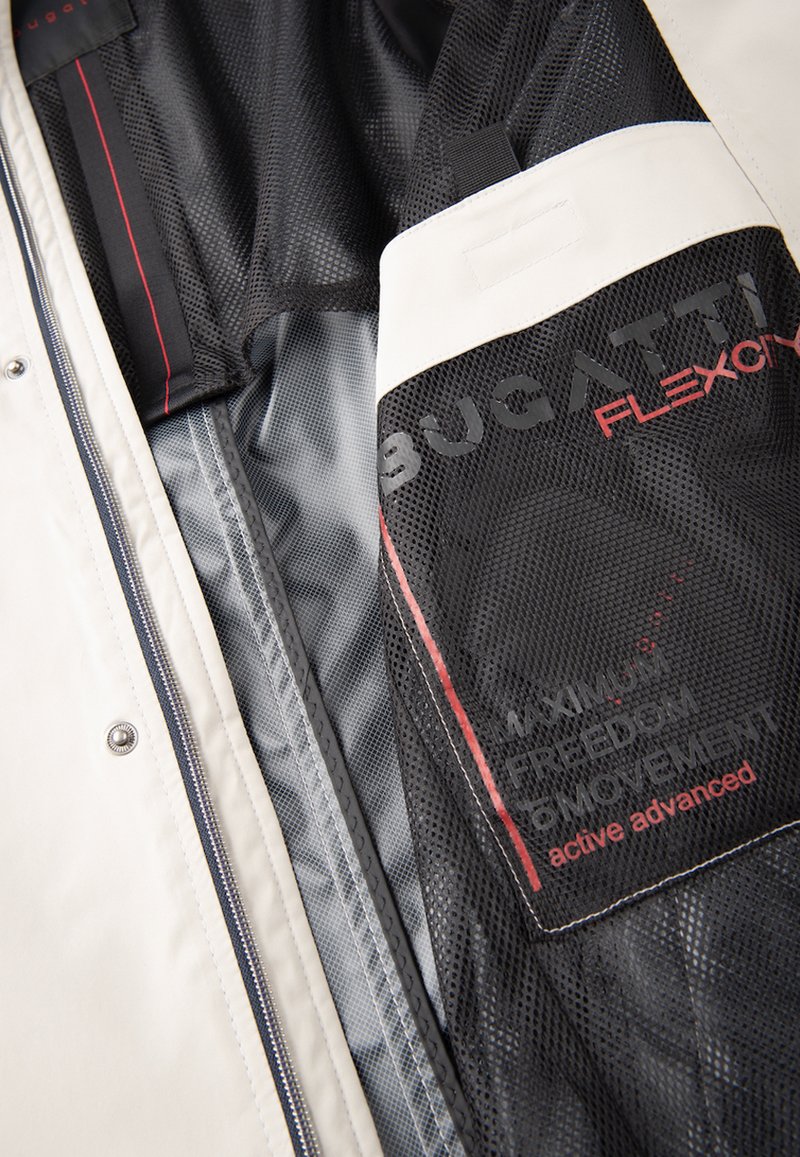 Intérieur de la veste blanche avec doublure en maille noire. Présente une étiquette avec un accent rouge indiquant "BUGATTI FLEXCITY." Poches zippées visibles.