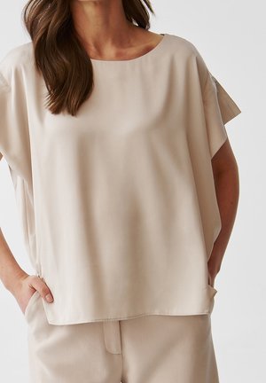 Frau trägt lockere beige Kurzarmbluse und passende Hose, Hände in den Taschen, vor schlicht hellem Hintergrund.