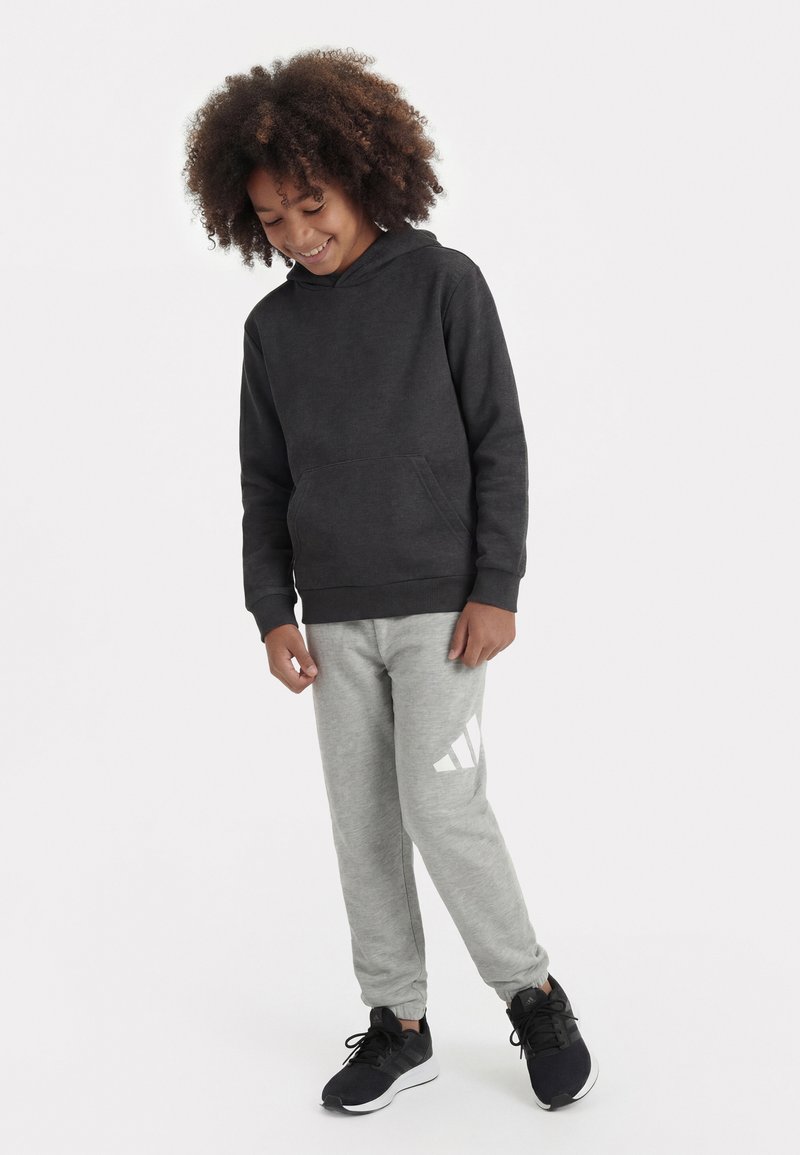 Enfant souriant aux cheveux bouclés portant un sweat à capuche noir, un pantalon de survêtement gris avec un logo blanc, et des baskets noires, debout devant un fond uni.