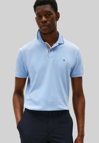 Homme portant un polo à manches courtes bleu clair et un pantalon bleu marine, les mains dans les poches, devant un fond gris uni.