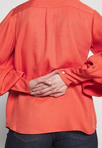 Blouse corail à manches longues avec poignets boutonnés et détail plissé dans le dos, en tissu lisse, associée à un jean en denim foncé.