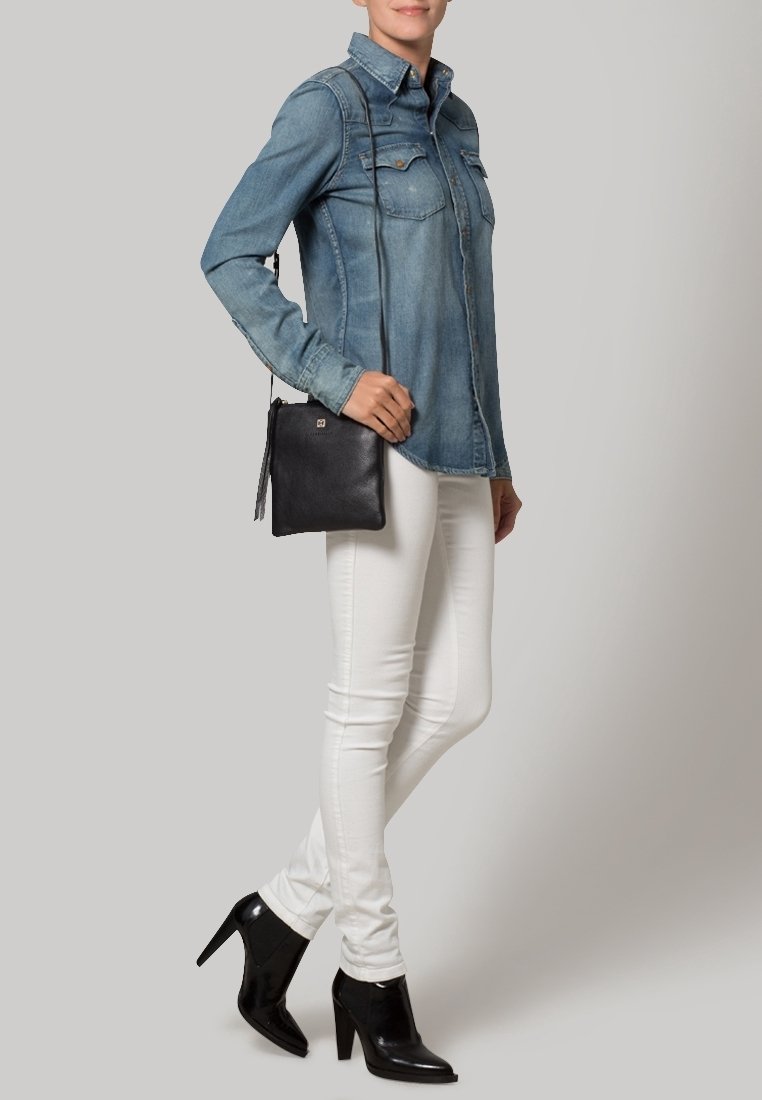 Musta nahkainen olkalaukku, jossa on teksturoitu pinta ja tupsukoriste, yhdistetty vaaleaan farkkupaitaan ja valkoisiin kapealahkeisiin farkkuihin.