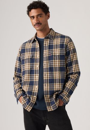 Mann mit lockigem Haar und Schnurrbart, der ein blau-beiges kariertes Hemd über einem dunkelblauen Pullover trägt und die Hände in den Taschen hat.