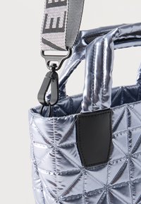 Γωνία καπιτονέ τσαντας ασημί μεταλλική που δείχνει επενδεδυμένες λαβές, μαύρο κούμπωμα και γκρι ρυθμιζόμενο λουρί με μαύρα γράμματα.