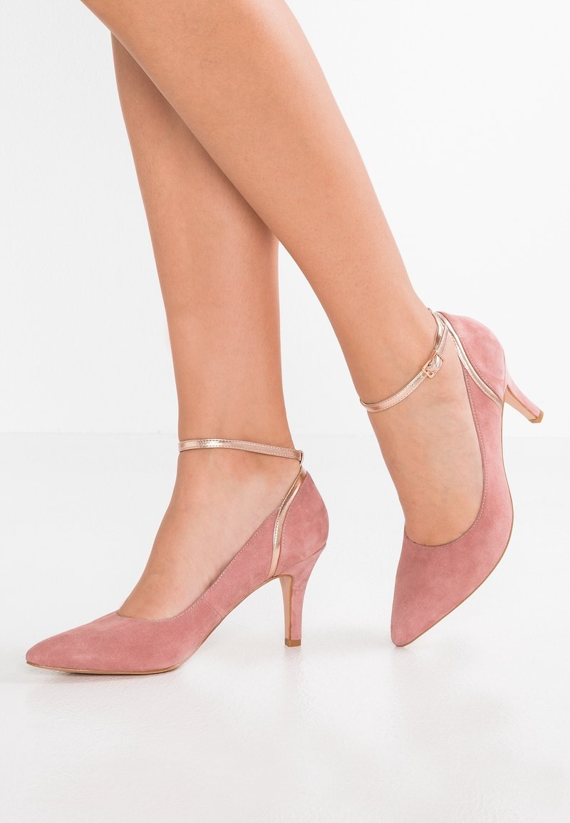 Jambes portant des escarpins en daim rose à bout pointu avec de fines brides métalliques à la cheville et des talons aiguilles de hauteur moyenne sur un sol blanc.