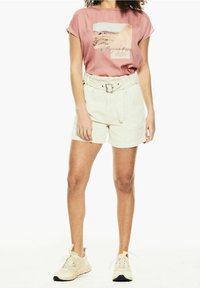 Růžové tričko s krátkým rukávem zdobí fotomotiv, spárované s béžovými šortkami s páskem. Modelka má na sobě světlé tenisky se žlutými akcenty.
