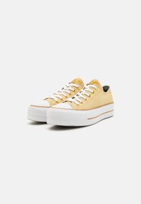 Zapatillas de lona amarillas con suelas de goma blanca, ojales dorados y forro a cuadros negros. Presenta una franja blanca sobre la suela.