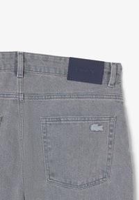Un jean en denim gris présente une étiquette en cuir bleu foncé avec "LACOSTE" en relief, ainsi qu'un petit logo de crocodile bleu clair sur la poche arrière.