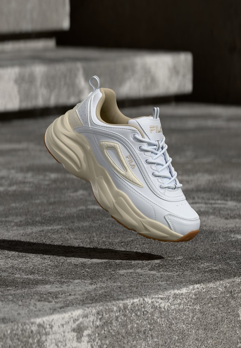 Λευκό και μπεζ αθλητικό παπούτσι FILA με χοντρή σόλα, που αιωρείται πάνω από υφασμένες σκάλες από σκυρόδεμα, ρίχνοντας σκιά από κάτω.