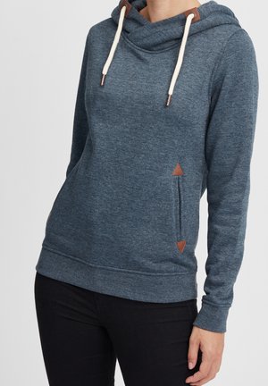 Personne portant un sweat à capuche bleu à manches longues avec des cordons blancs et des détails en cuir marron, associé à un pantalon noir.