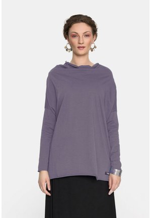 Kobieta o kręconych włosach, ubrana w luźną lawendową bluzkę z długim rękawem, duże kolczyki koła w kształcie serca, srebrną bransoletkę i czarną spódnicę.