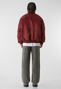 Burgundská bomber bunda s elastickými manžetami a lemem, v kombinaci se šedými širokými džínami s zadními kapsami a jednoduchým bílým tričkem pod ní.